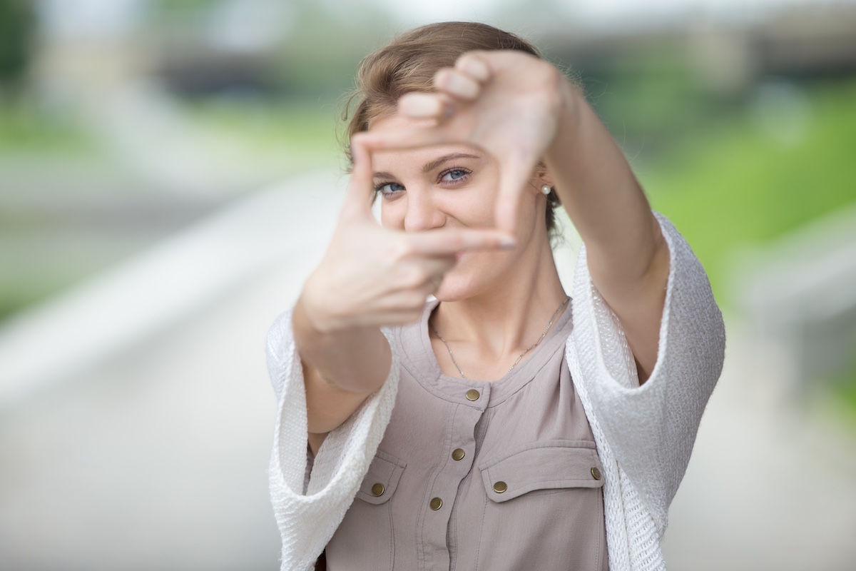 Portrait of playful beautiful caucasian woman pretending to take photo with her hands while walking on the street. Attractive model posing using hands to make a frame outdoors in summer
